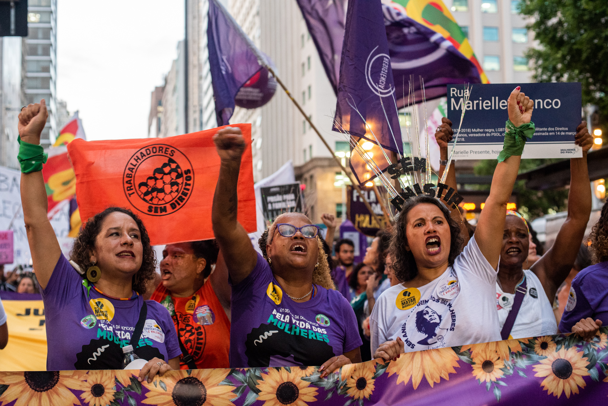 Women at the beginning of the 8M act hold a banner and shout slogans. In the photo, from left to right: Councilor Luciana Boiteux, Councilor Monica Cunha, Tatianny Araújo; in the back, holding the flag of the Workers Without Rights Movement: Maria dos Camelôs and, holding Marielle Franco’s plaque, Sílvia Mendonça. Photo: Bárbara Dias